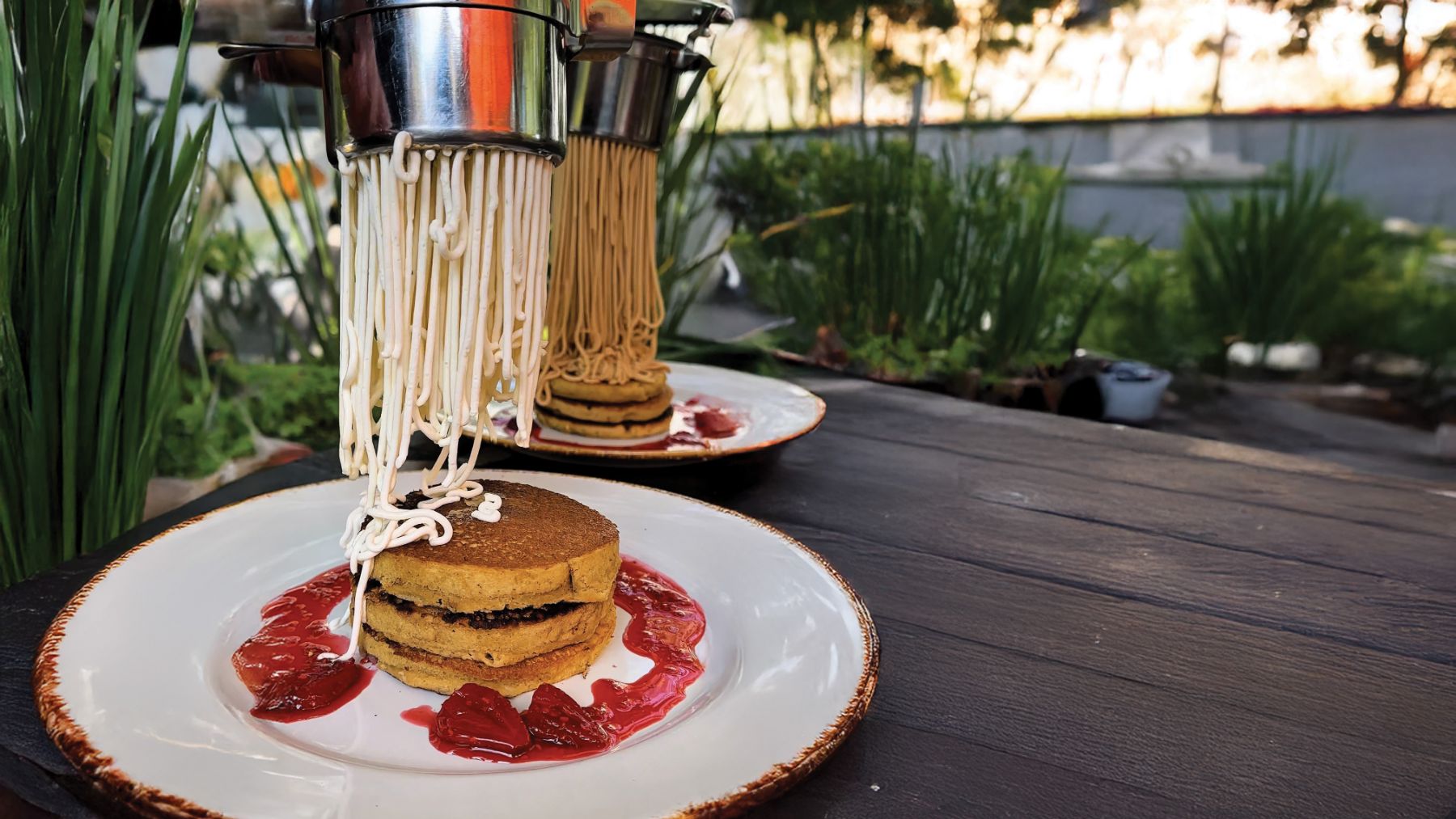 La prensa de los Pancakes Spaguetti deja caer hilos de crema sobre una torre de pancakes con frutos rojos, en una mesa del jardín