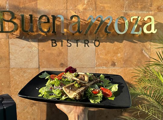 El letrero de Buena Moza Bistró en latón sobre un muro de piedra, con una ensalada en primer plano